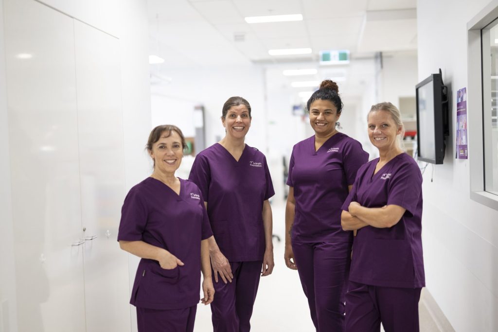 Four nurses in purple scrubs stand in a hospital hallway, smiling.