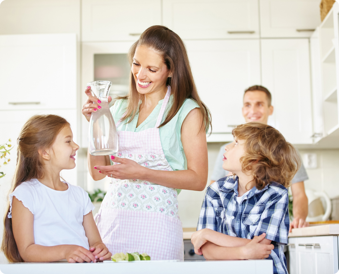 A mother pours water from a carafe for her children in a bright kitchen.