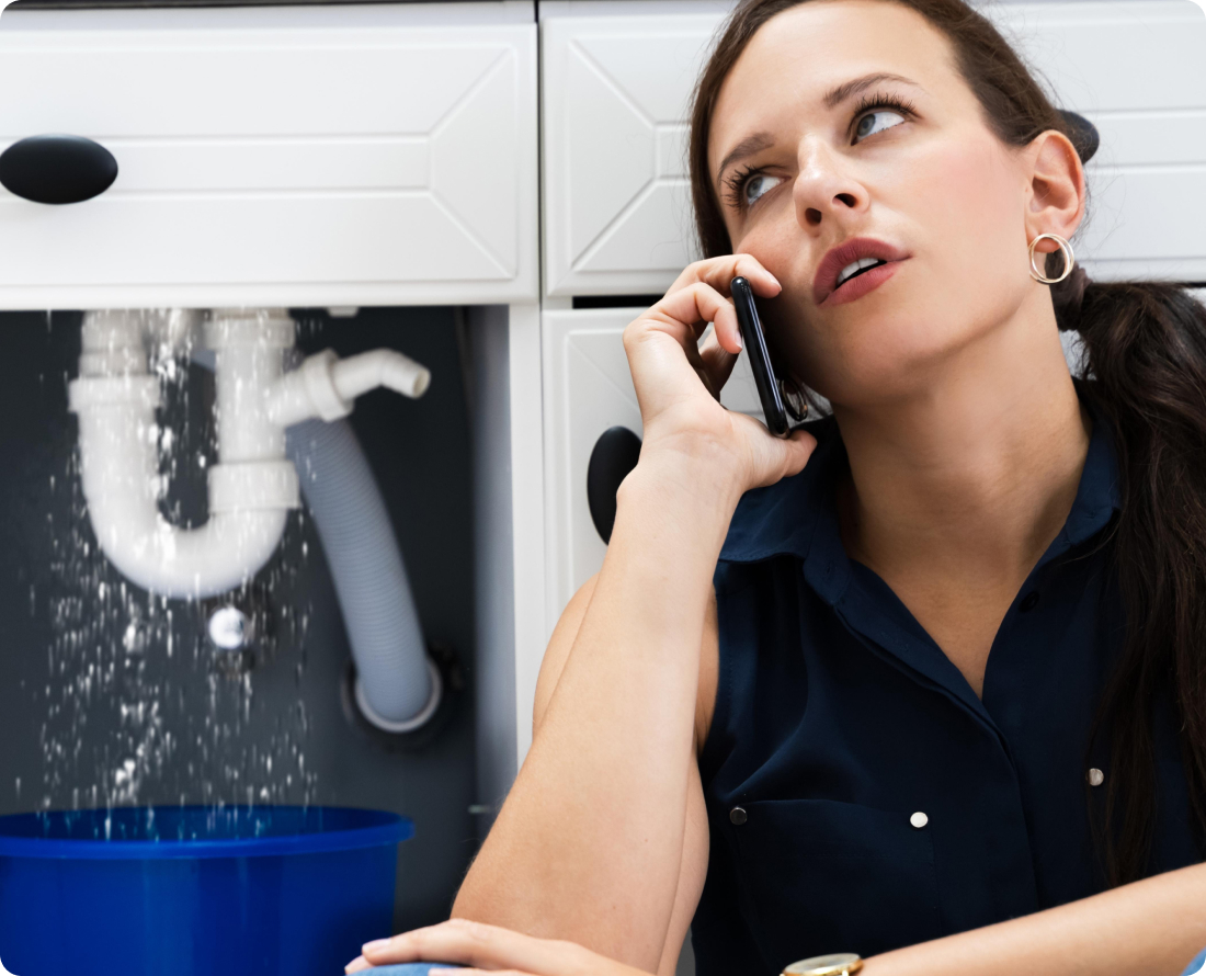 Woman on phone by leaky sink, water collecting in blue bucket.