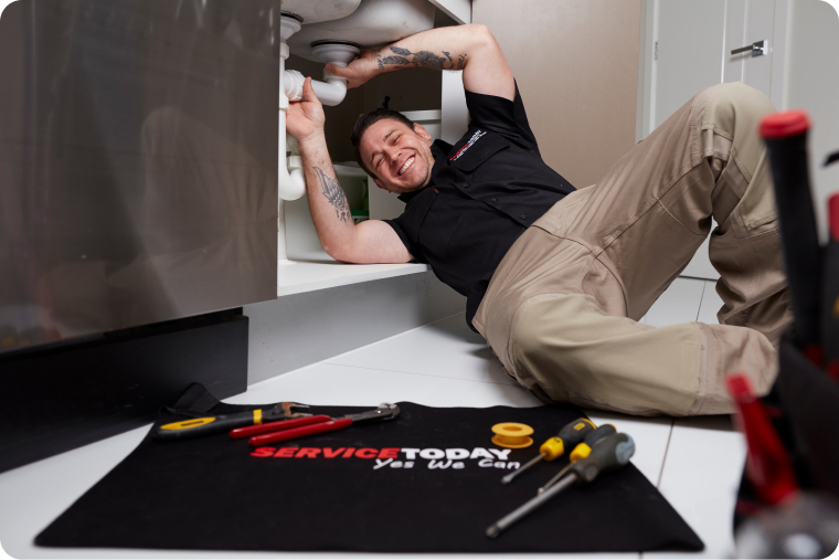 Plumber working on pipes under a sink, with "Service Today" mat and tools.