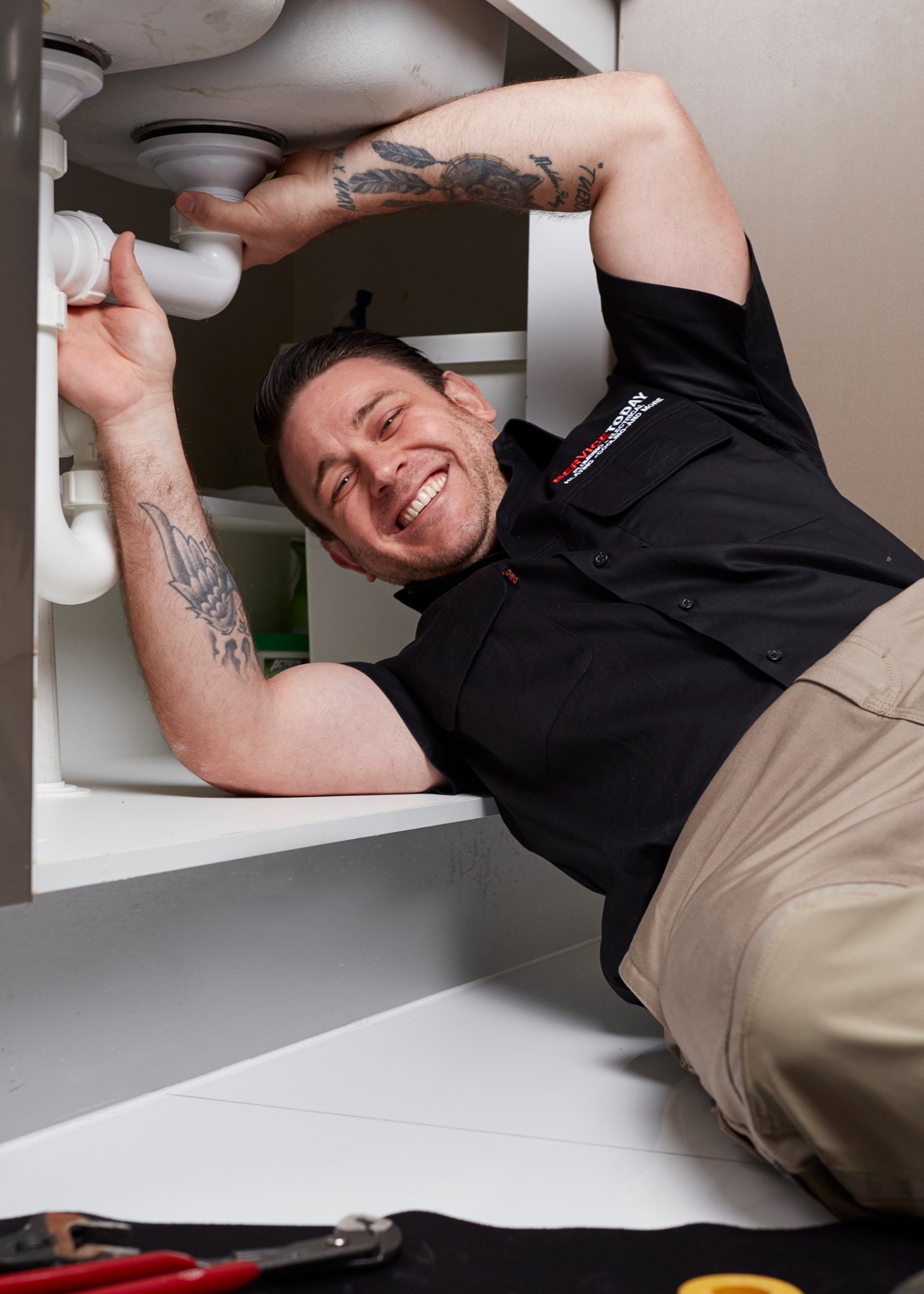 Plumber smiling while fixing pipes under a sink, wearing a black shirt and tan pants.