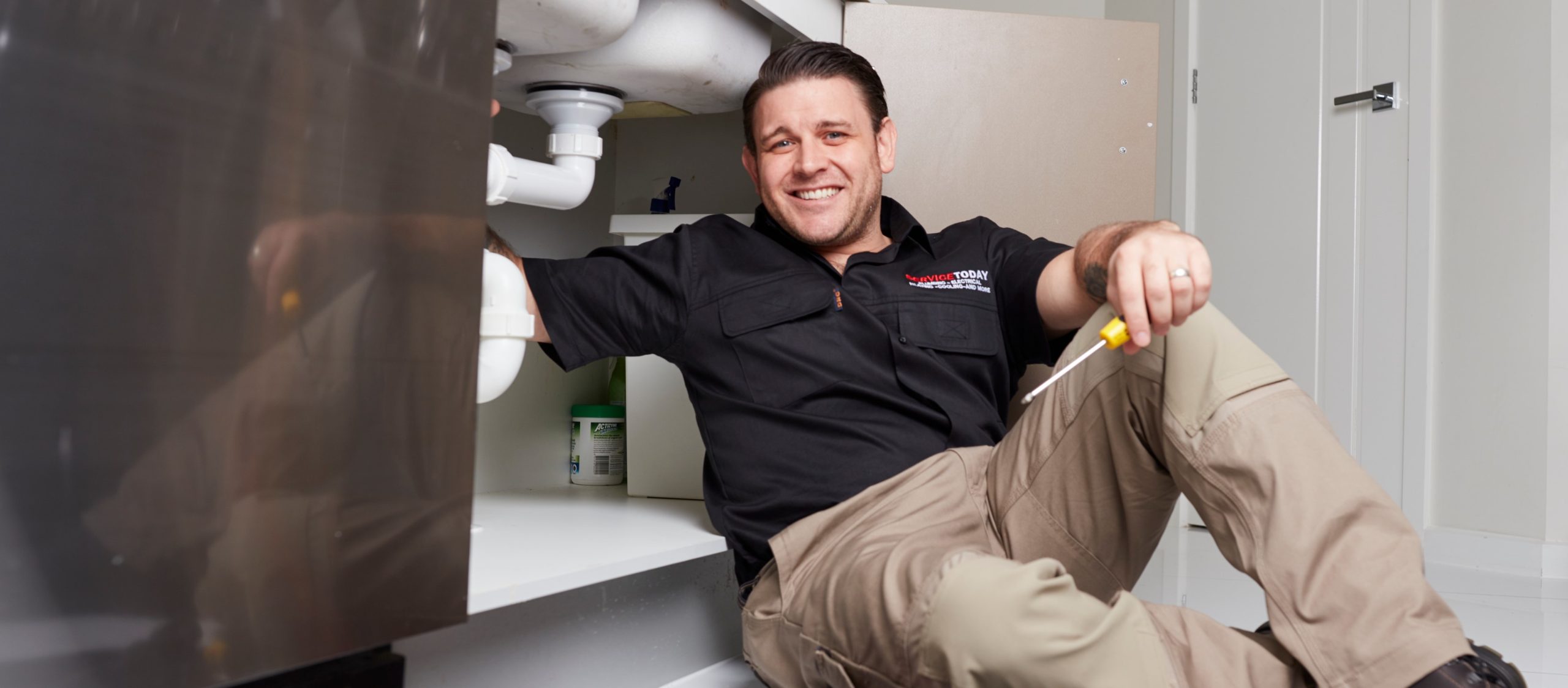 Smiling plumber in black shirt and tan pants works under a sink, holding a screwdriver.