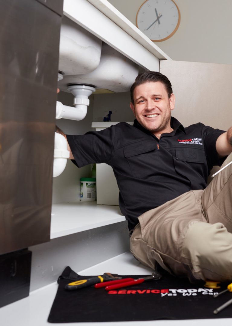 Plumber smiling while fixing a sink with tools and cleaning supplies nearby.
