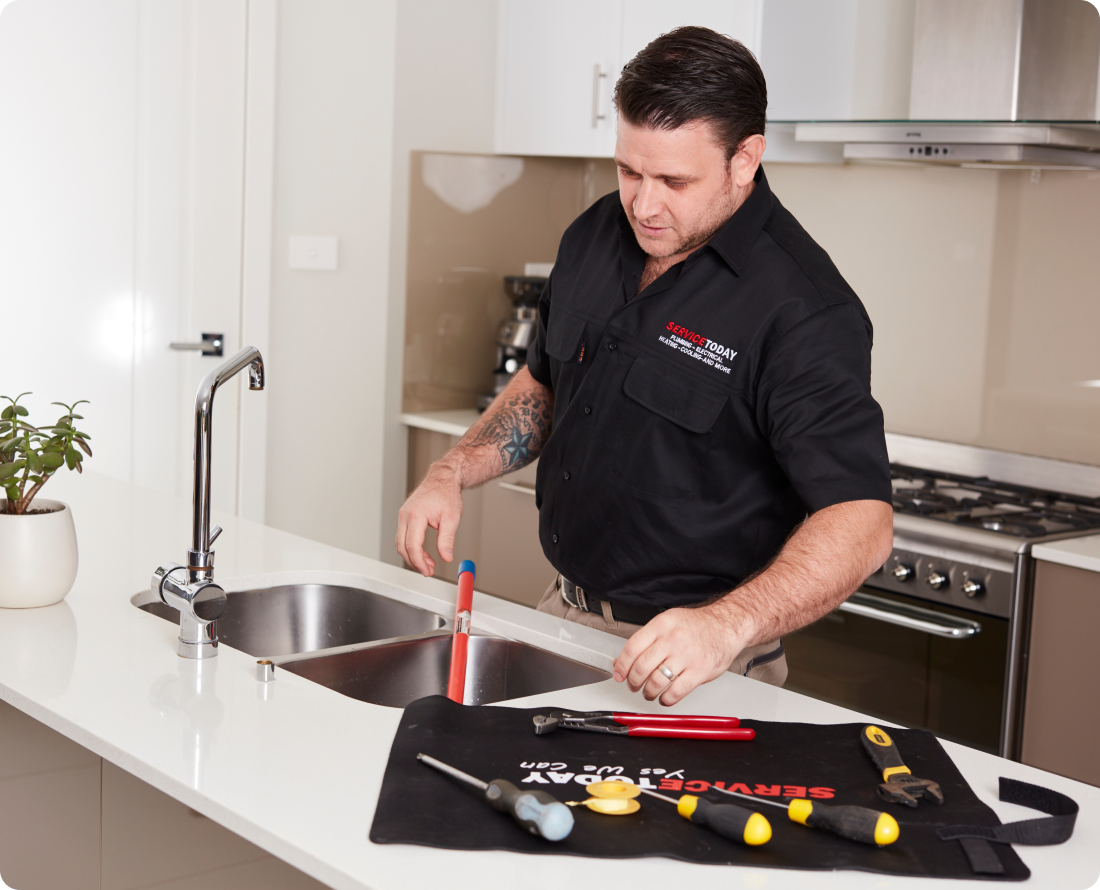 Plumber at kitchen sink fixing faucet with tools on mat.