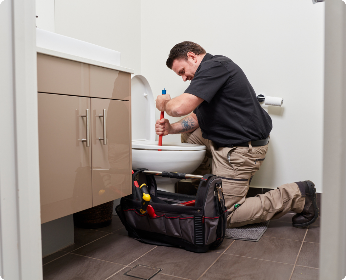 Plumber unblocking a toilet with a plunger, with a toolbox nearby.