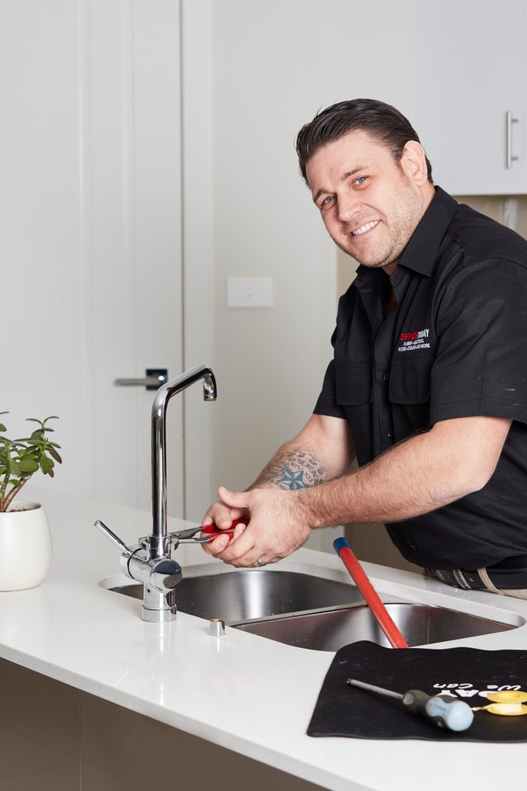 Plumber fixing a kitchen tap with red pliers, smiling at the camera.